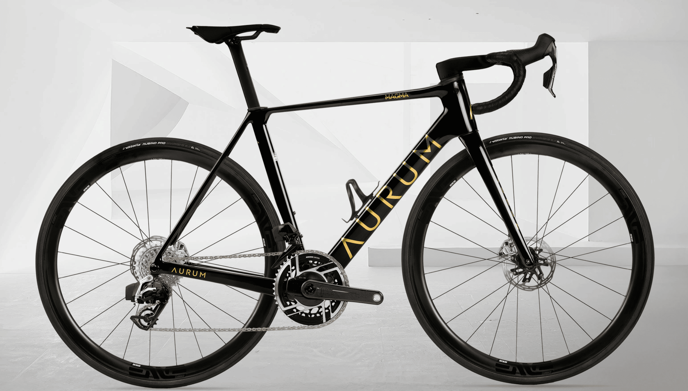 Sort AURUM Magma landeveissykkel med gull detaljer og ENVE-hjul, fotografert fra siden mot en lys, hvit bakgrunn med geometriske former.