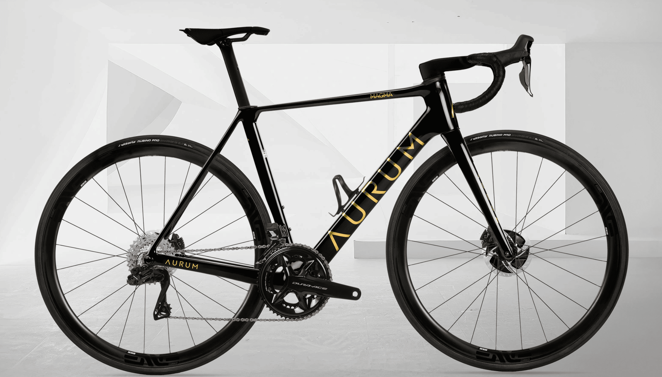 Sort AURUM Magma landeveissykkel med gullfargede detaljer og ENVE-hjul, fotografert fra siden mot en lys, geometrisk hvit bakgrunn.